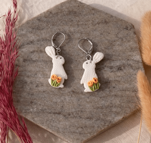 Handmodellierte Hasen Ohrringe aus weißem Polymerton mit roten Rosen und hypoallergenem Edelstahl-Verschluss auf Naturstein fotografiert