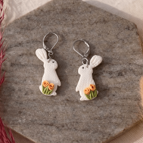 Handmodellierte Hasen Ohrringe aus weißem Polymerton mit roten Rosen und hypoallergenem Edelstahl-Verschluss auf Naturstein fotografiert